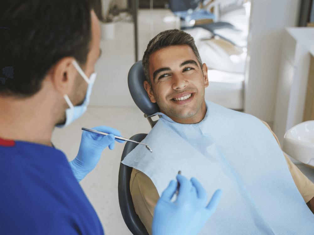 A patient with a blue bib on his chest smiling to the dentist. The dentist hold a dental instrument, in a dental setting.