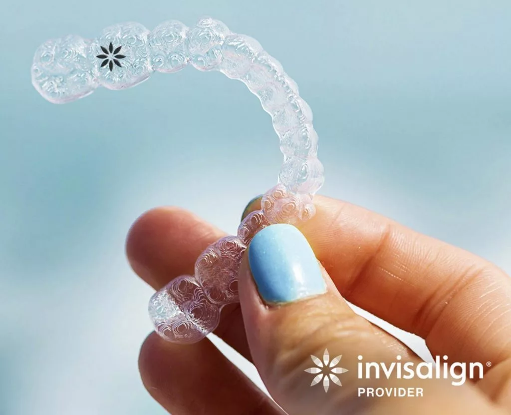 Close-up of a clear dental aligner held by a hand with light blue painted nails, featuring the Invisalign logo in the bottom right corner.