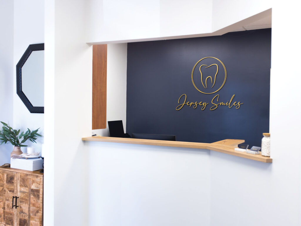 Reception area of a dental office named 'Jersey Smiles,' featuring a modern wooden desk, a dark blue wall with a gold tooth logo and the business name written in cursive, along with stylish décor including a plant, hexagonal mirror, and wooden cabinet.
