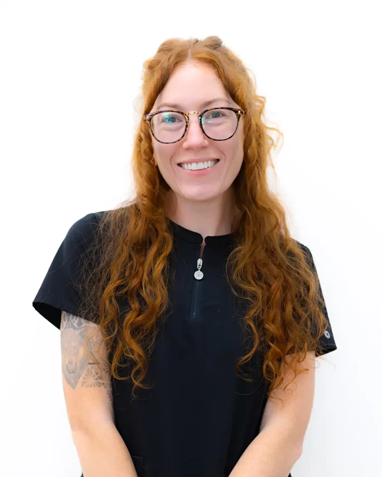 Brianna, a dental hygienist, is in a portrait against a plain white background with long wavy red hair, glasses, a short‑sleeved black shirt, and visible arm tattoos.