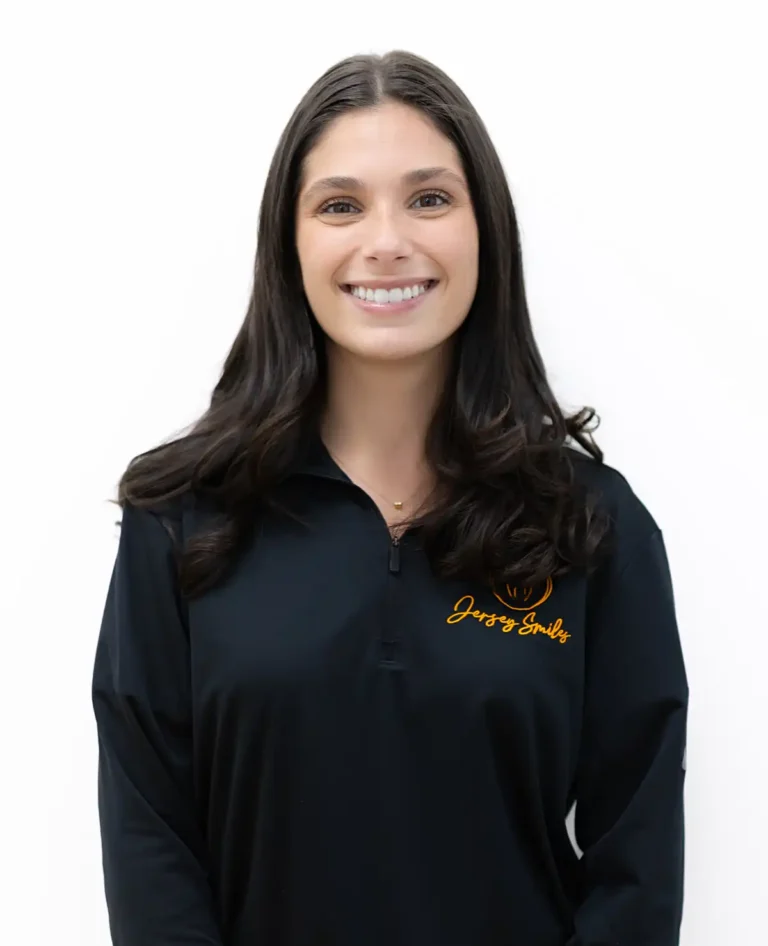 Samantha, front office administrator at Jersey Smiles, professional portrait against plain white background wearing black quarter‑zip pullover with orange embroidered logo.