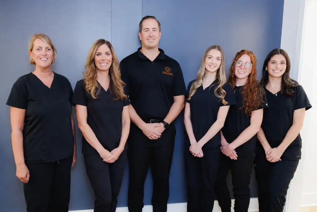 Group photo of Jersey Smiles dental team standing side by side in front of blue wall, all wearing black professional attire including scrubs and polo shirt with orange logo, posed formally for staff portrait.