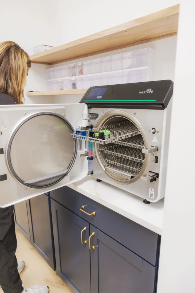 Person standing indoors beside open sterilization autoclave built into cabinet, with visible metal racks inside and surrounding clinical workspace.