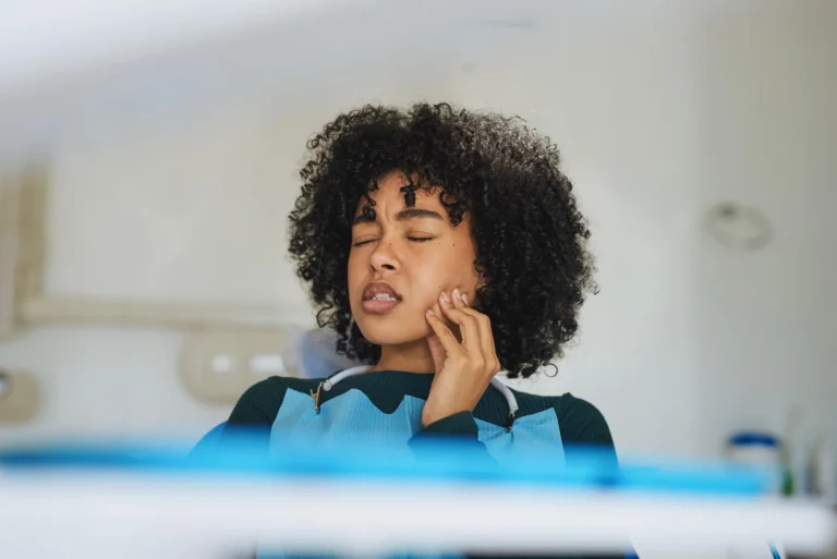 A woman with curly hair pressing a hand against the side of their jaw with a pained expression due to TMJ disorder
